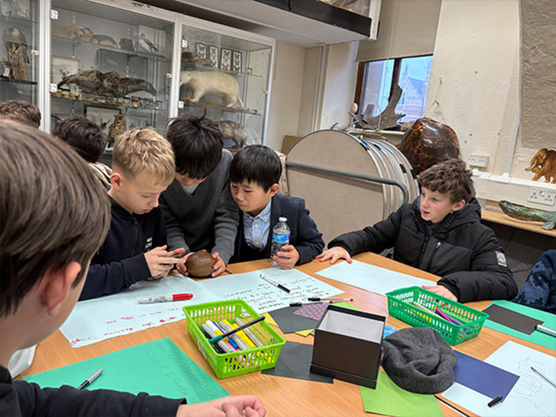 Several students sit around a table covered with colored paper, markers, and supplies, closely examining a small brown object together while others watch and participate.