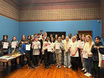A large group of students and adults pose together in a classroom, many holding certificates and smiling toward the camera.