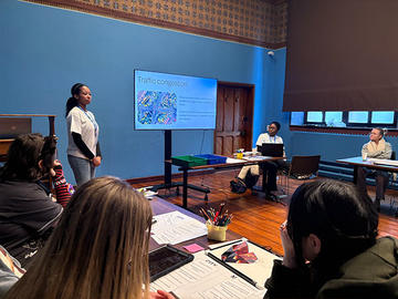 Two students stand beside a screen displaying the UN Sustainable Development Goals wheel, with one pointing to the graphic while presenting.