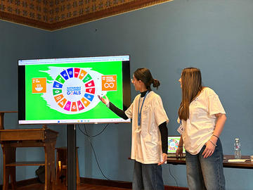 Two students stand beside a screen displaying the UN Sustainable Development Goals wheel, with one pointing to the graphic while presenting.